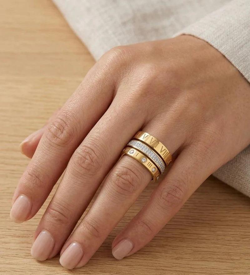 A close-up of a hand wearing three stacked rings on the ring finger. The rings include a yellow gold band engraved with letters, a silver band with small clear stones, and a yellow gold band with initials.