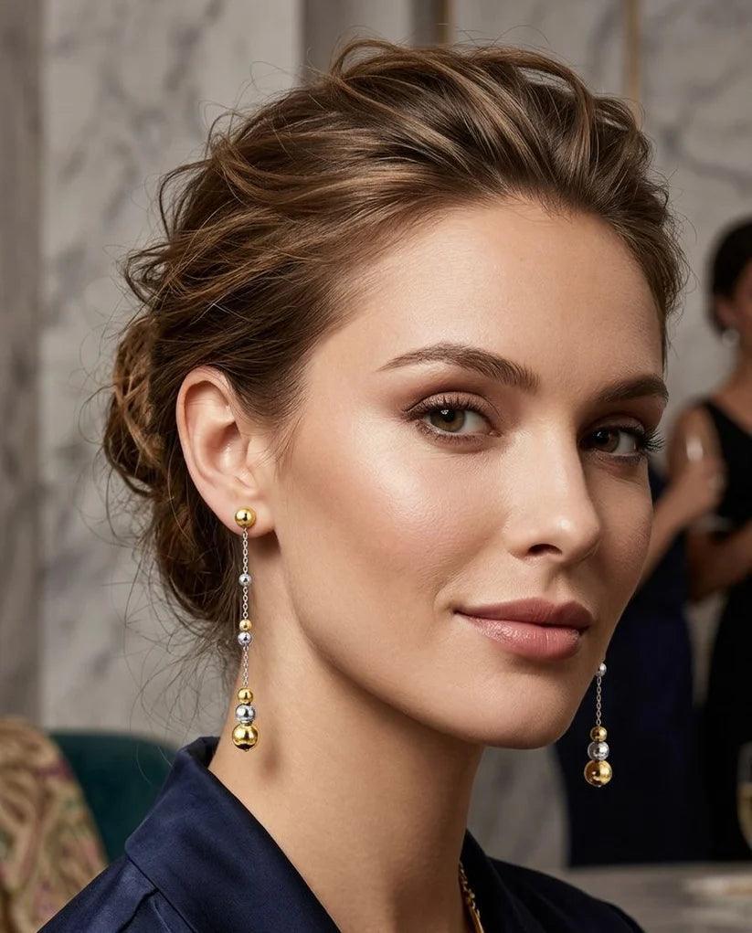 A close-up portrait of a woman wearing elegant gold and silver drop earrings with small beads and larger gold ball accents, set against a softly blurred indoor background.