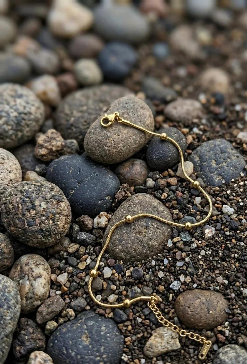 A delicate gold chain bracelet with small bead accents and a lobster clasp, laid out on a bed of smooth river stones and gravel.