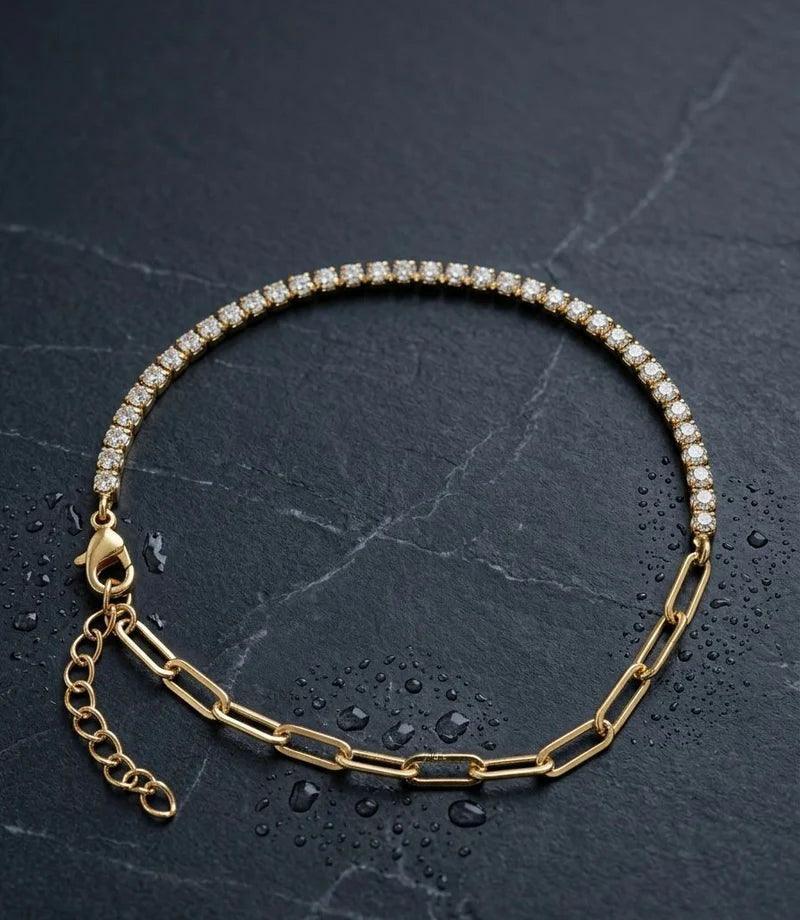 A gold tennis bracelet with a row of clear stones on one side and paperclip links on the other, displayed on a dark textured slate surface with water droplets.