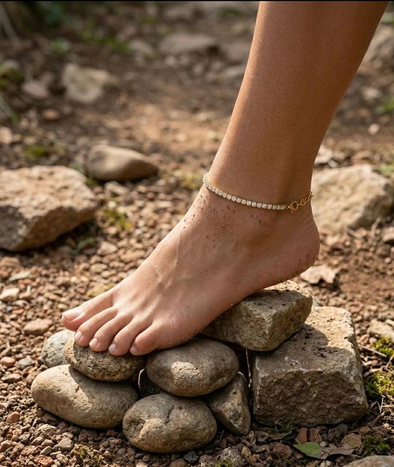 A close-up of a bare foot resting on stacked stones outdoors, wearing a delicate gold anklet with small clear stones.