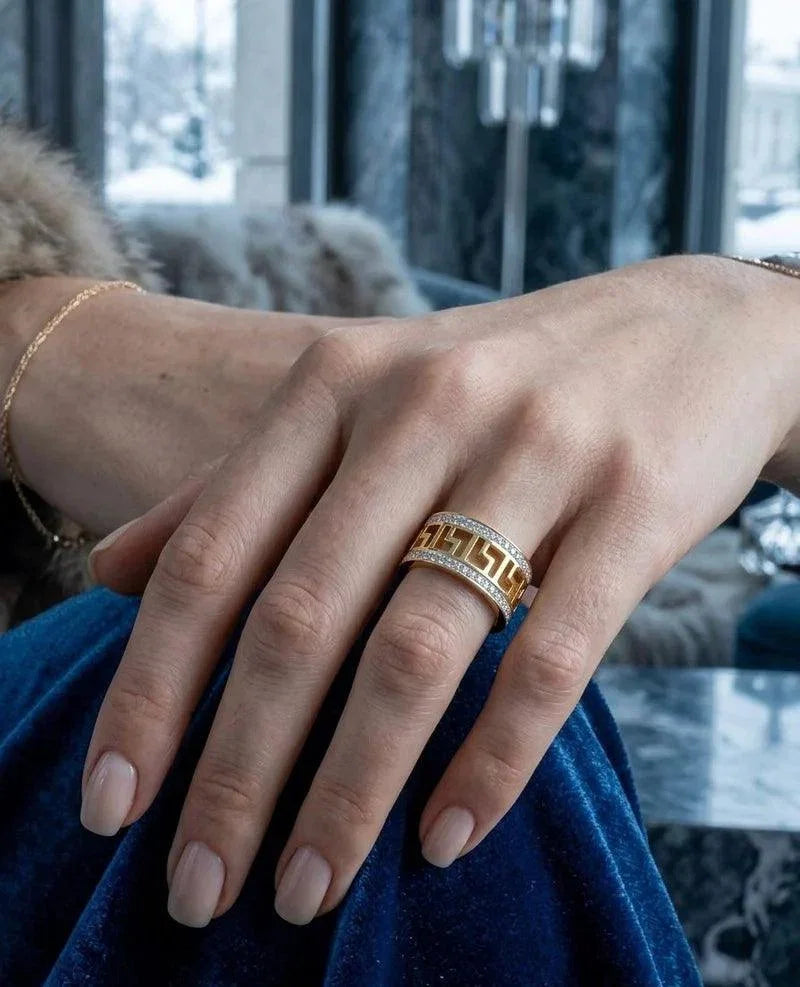 A close-up of a hand wearing a gold ring with a Greek key pattern and a row of small clear stones, resting on blue velvet fabric in an indoor setting.