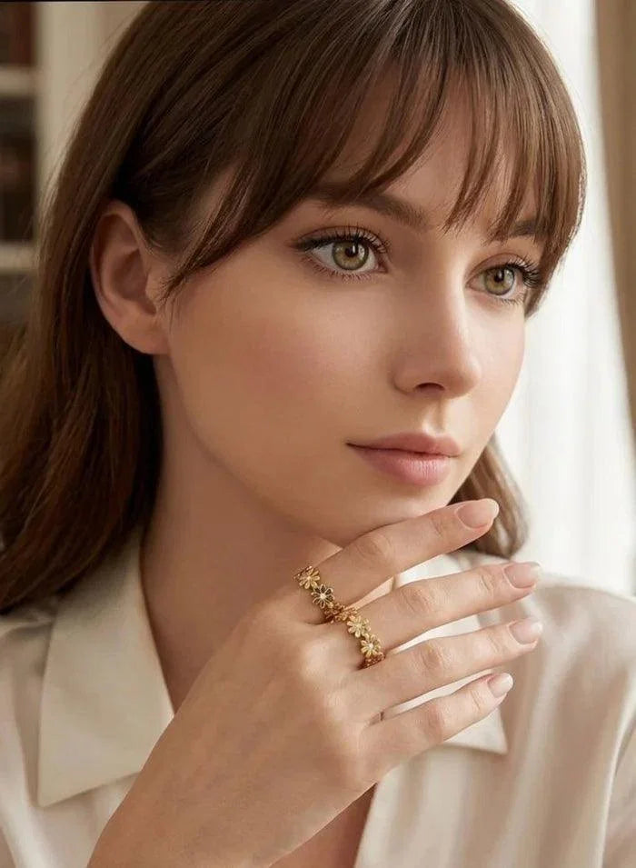 A close-up portrait of a woman with brown hair and bangs wearing a delicate gold ring with a floral or vine-like pattern on one finger. She is wearing a light cream-colored collared shirt, and the background is a softly blurred indoor setting with warm light.