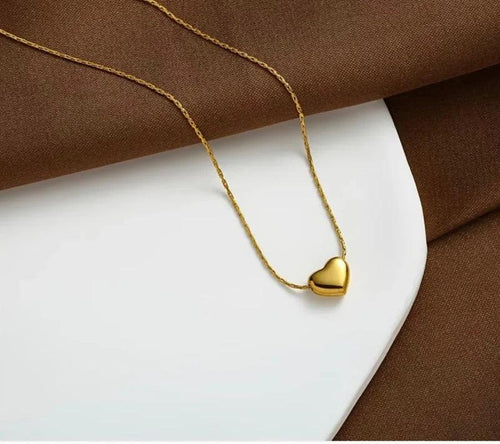 A delicate gold chain necklace with a small, polished gold heart pendant, styled on a white plate with brown fabric in the background.