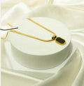A gold chain necklace with a geometric pendant featuring a black center and gold border, displayed on a white cylindrical pedestal against a draped white satin background.