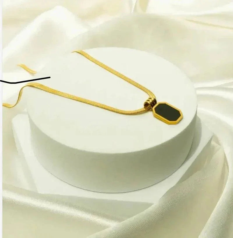 A gold chain necklace with a geometric pendant featuring a black center and gold border, displayed on a white cylindrical pedestal against a draped white satin background.