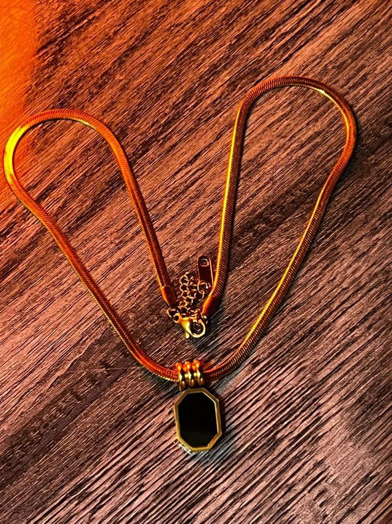 A gold-toned snake chain necklace with a black faceted octagonal pendant, displayed on a textured brown wooden surface.
