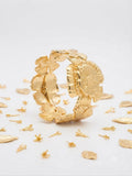 A close-up of a gold floral cuff bracelet worn on a person's wrist, with their hand resting gently above it against a soft neutral background.