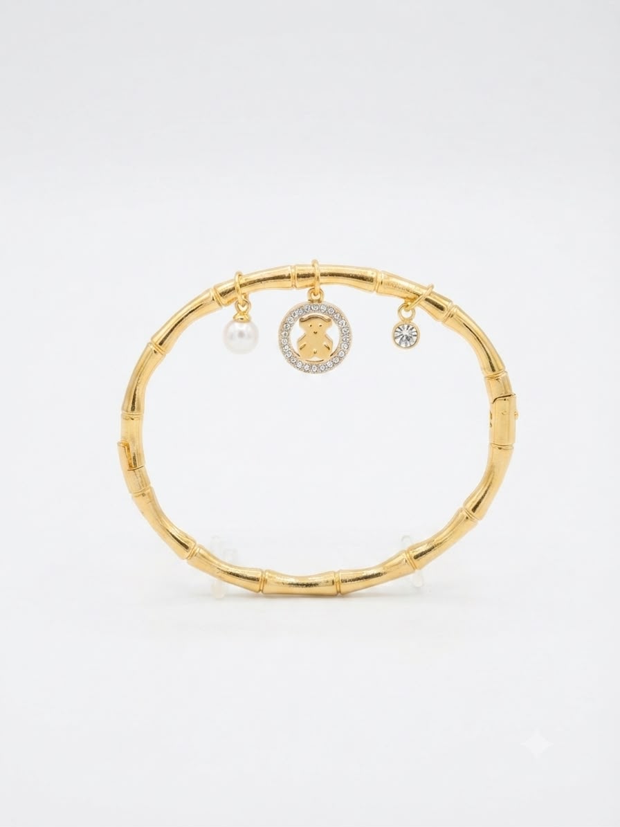 A gold-toned bangle bracelet with a bamboo-inspired segmented design, featuring three hanging charms: a white pearl, a round gold charm with a teddy bear motif surrounded by small clear stones, and a small clear stone charm, set against a plain white background.