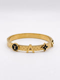 A gold-tone Louis Vuitton bangle bracelet featuring the brand's monogram pattern, a circular LV logo medallion, a large letter 'A' charm, and a black enamel flower accent, shown on a light gray background.