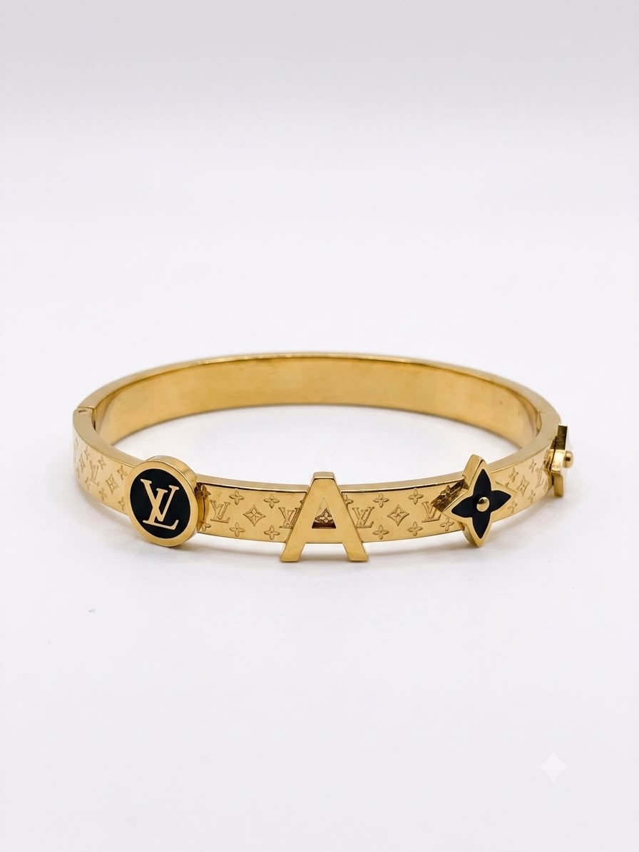 A gold-tone Louis Vuitton bangle bracelet featuring the brand's monogram pattern, a circular LV logo medallion, a large letter 'A' charm, and a black enamel flower accent, shown on a light gray background.