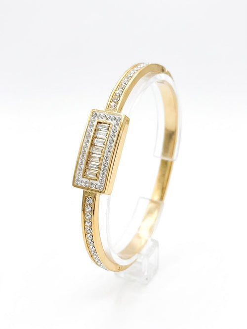 A gold bangle bracelet with a rectangular bezel-set diamond centerpiece and rows of small diamonds along the band, displayed on a clear stand against a white background.