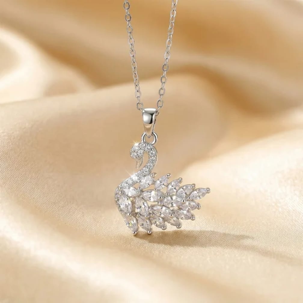A silver-toned swan pendant necklace adorned with clear crystals, resting on draped beige satin fabric.