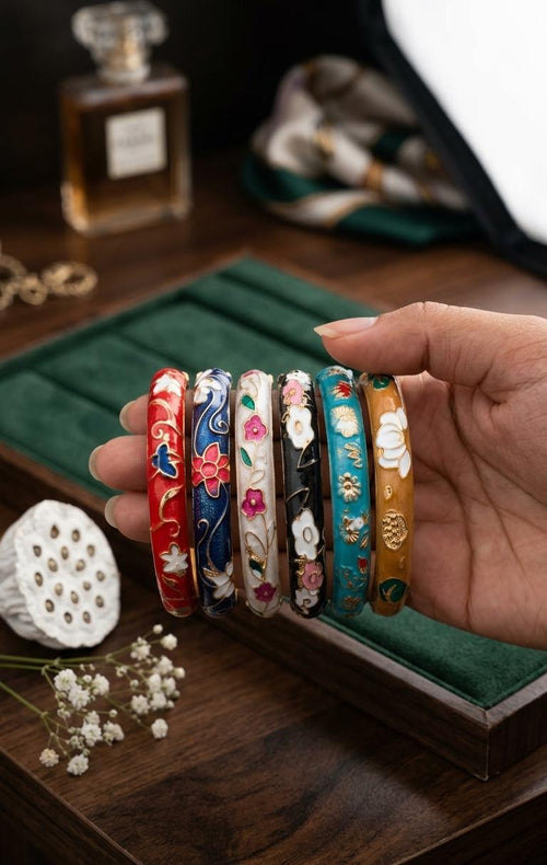 A hand holds six colorful, hand-painted Chinese bangles with floral engravings over a green jewelry display tray on a wooden table.