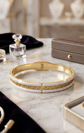 A gold and white bangle bracelet with a Greek key pattern, displayed on a marble surface with luxury accessories in a soft-focus background.