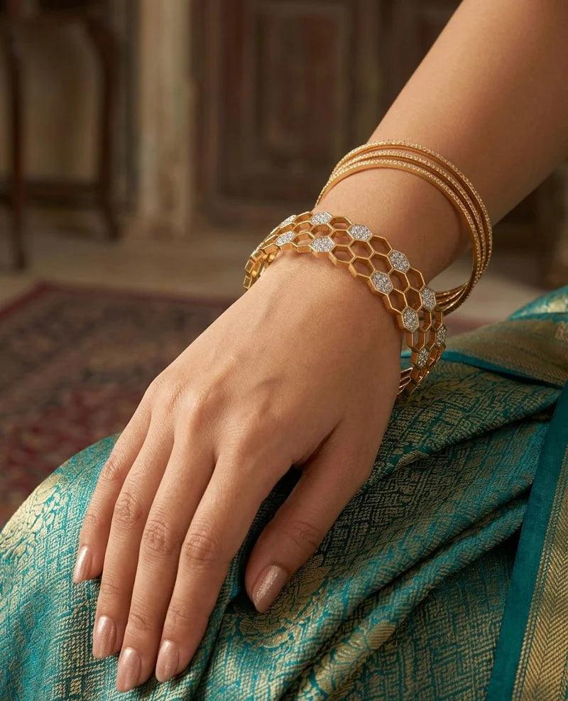 A close-up of a woman's hand and wrist wearing a stack of gold bangles, including a prominent honeycomb-style bracelet with clear stones, resting on her teal and gold patterned sari.