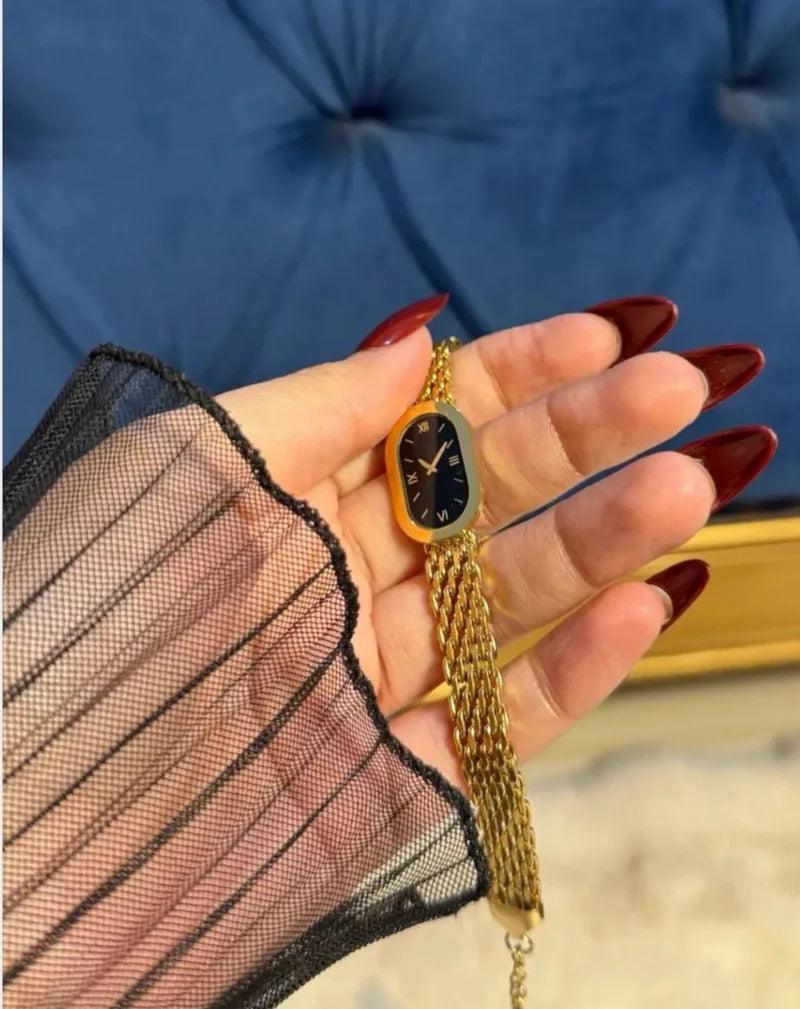 Hand holding a gold chain necklace with a black pendant against a blue background