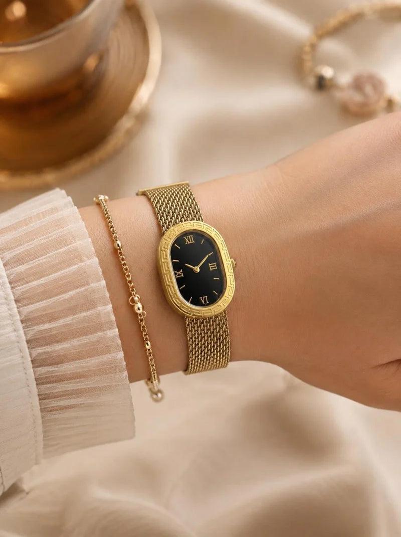 A gold-tone wristwatch with an oval black dial and mesh band, shown worn on a person's wrist alongside a delicate gold chain bracelet, set against a soft beige fabric background.