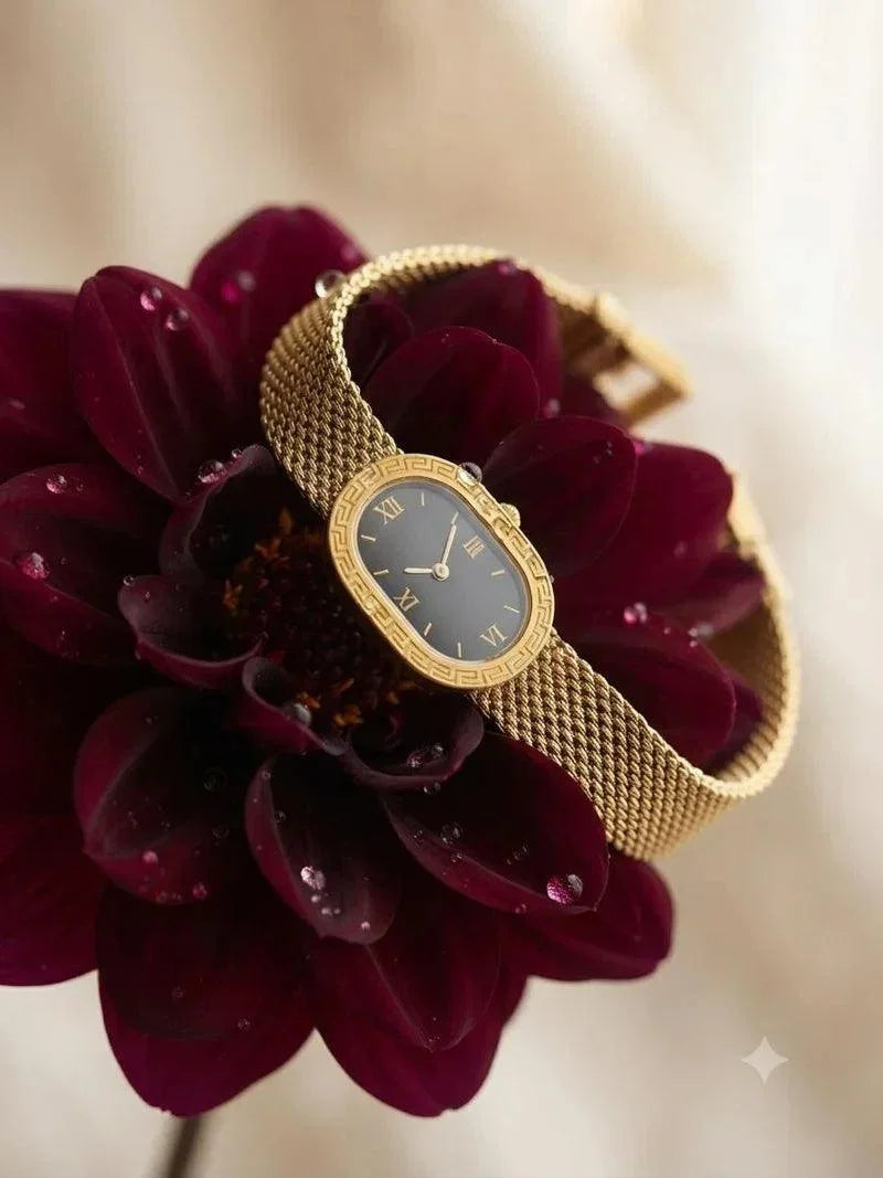 A gold-tone mesh-band wristwatch with a dark dial and gold-tone bezel, artfully draped over a deep burgundy flower against a soft cream background.