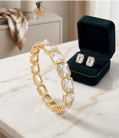 A gold bracelet with clear rectangular gemstones is displayed on a marble surface next to an open black jewelry box containing matching stud earrings.