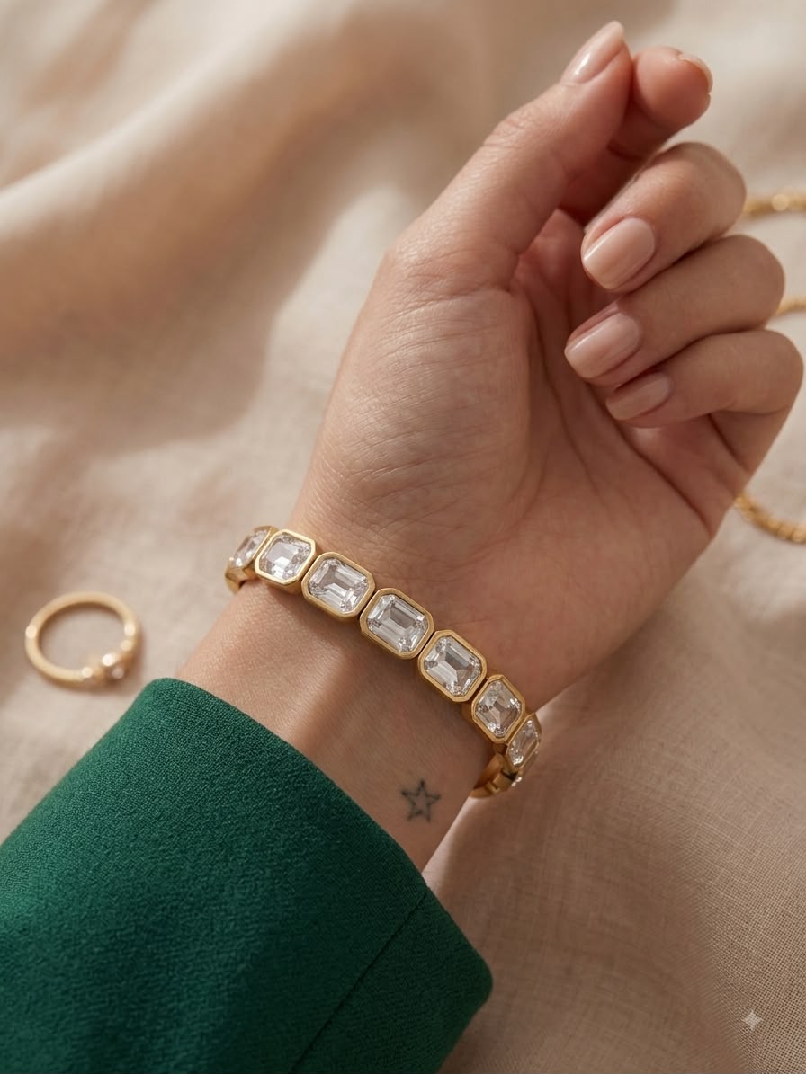 A gold bracelet with rectangular clear crystals worn on a woman's wrist, shown against a soft beige fabric background with a green sleeve visible.