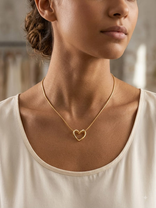 A close-up of a woman wearing a delicate gold chain necklace with a small open heart pendant, paired with a cream-colored scoop-neck top.