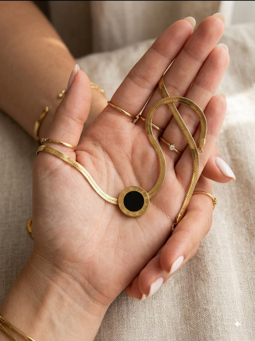 A close-up of a person's hands holding a gold snake chain necklace with a round black inlay, styled with matching gold rings and bracelets.