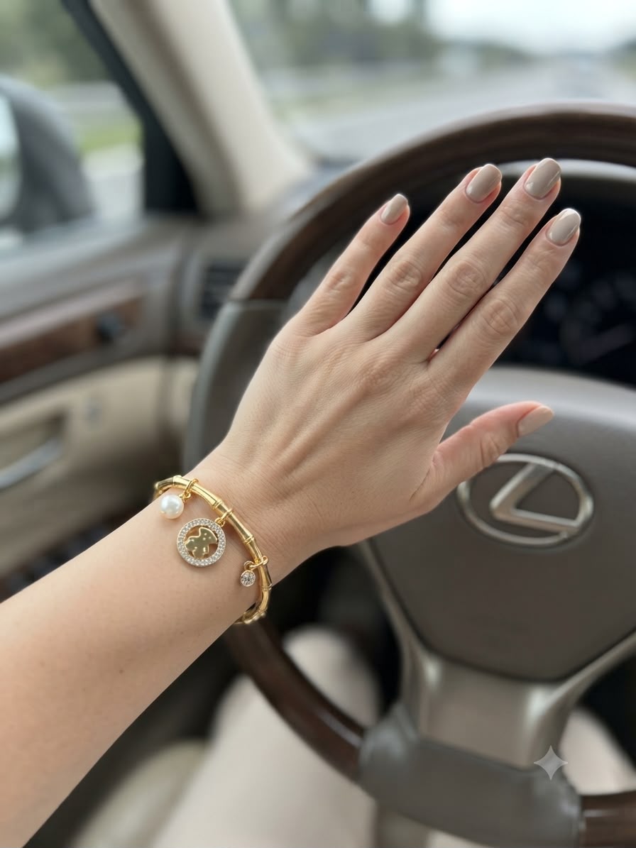 A gold-toned bangle bracelet with a round bezel-set stone and a small pearl accent, worn on a person's wrist inside a car with a steering wheel visible in the background.