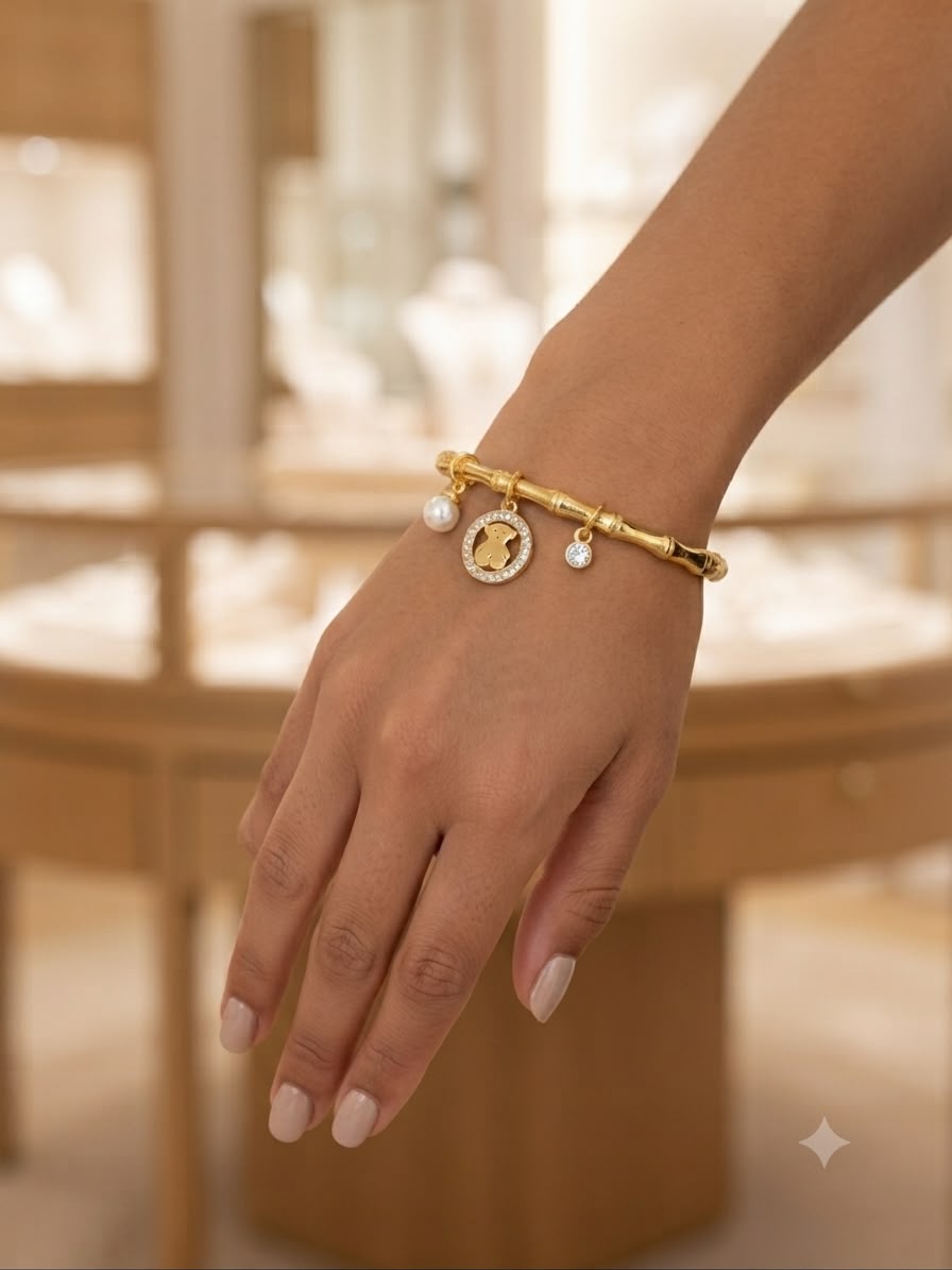 A gold charm bracelet with a bamboo-inspired link design, a central oval charm, and two small pearl-like beads, shown worn on a person's wrist in a softly blurred jewelry store setting.