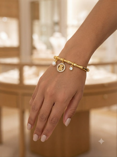 A gold charm bracelet with a bamboo-inspired link design, a central oval charm, and two small pearl-like beads, shown worn on a person's wrist in a softly blurred jewelry store setting.