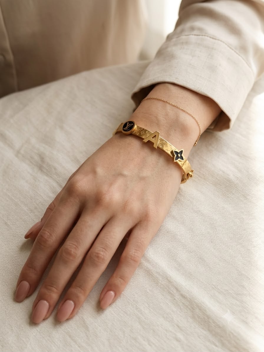 A close-up of a person's hand and wrist wearing a gold-tone bracelet with star and oval motifs, along with a delicate chain bracelet, resting on a light beige fabric surface.