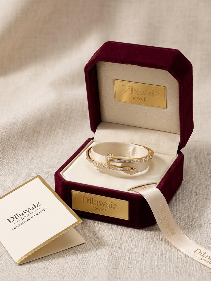 A gold-toned bangle bracelet with clear pavé stones, displayed inside an open burgundy velvet jewelry box with a gold plaque. A small branded card is placed beside the box on a light fabric surface.