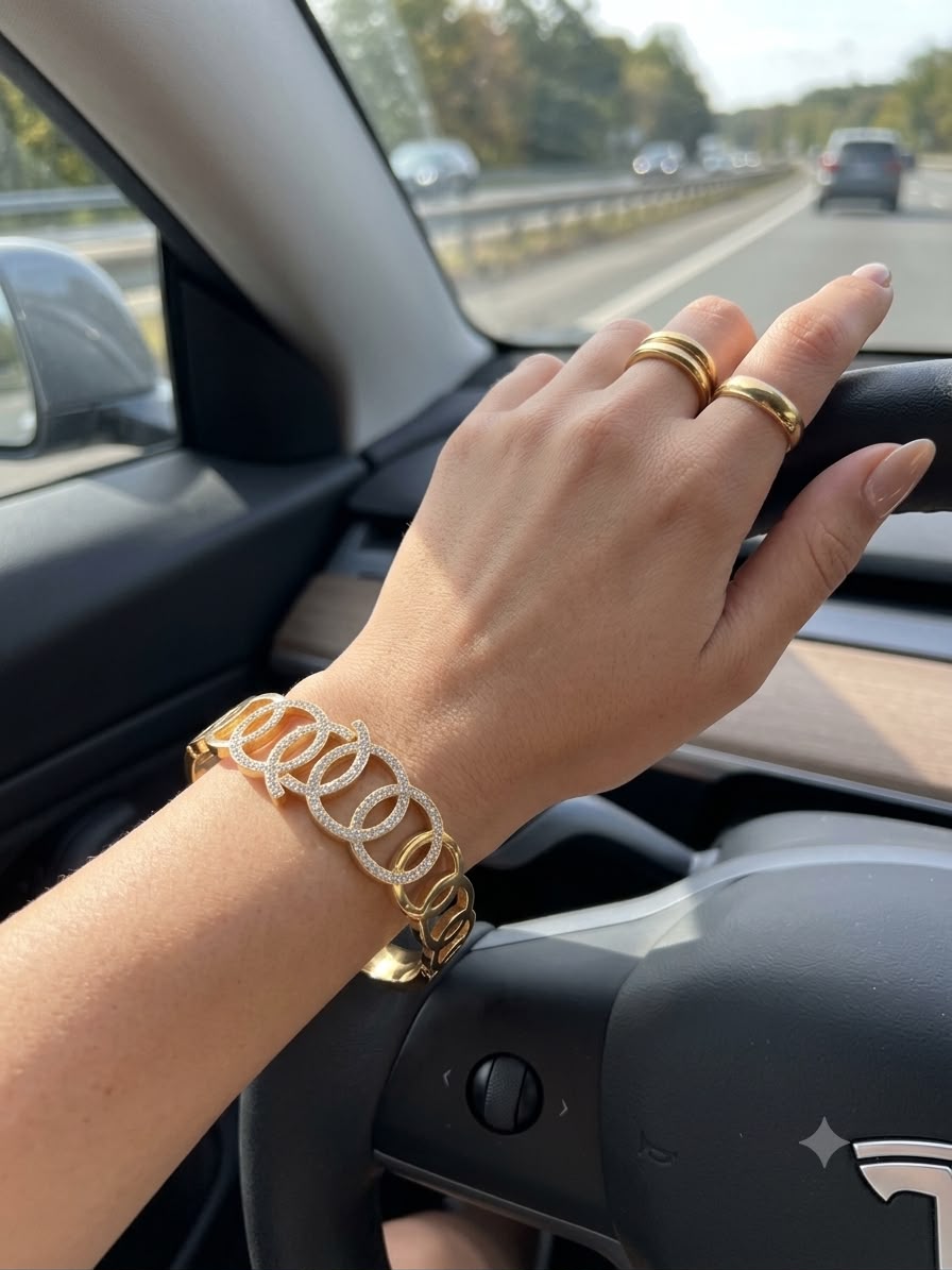 A gold chain bracelet with clear rhinestone links worn on a person's wrist inside a car, with the hand resting on the steering wheel.