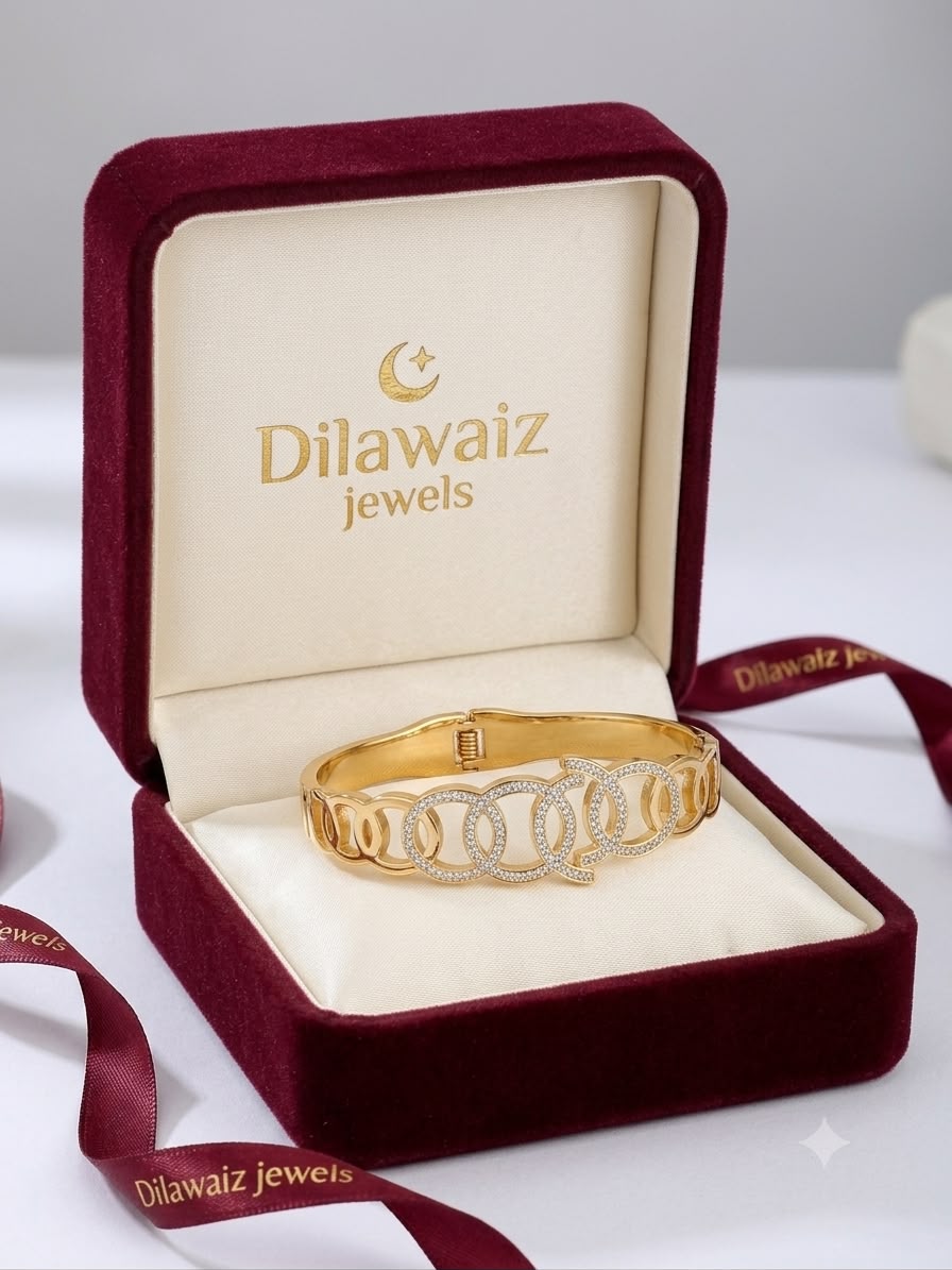 A gold bangle bracelet with a crescent moon and stars motif, displayed inside an open burgundy velvet jewelry box with cream lining. The inside lid features the brand name 'Dilawaiz jewels' in gold.