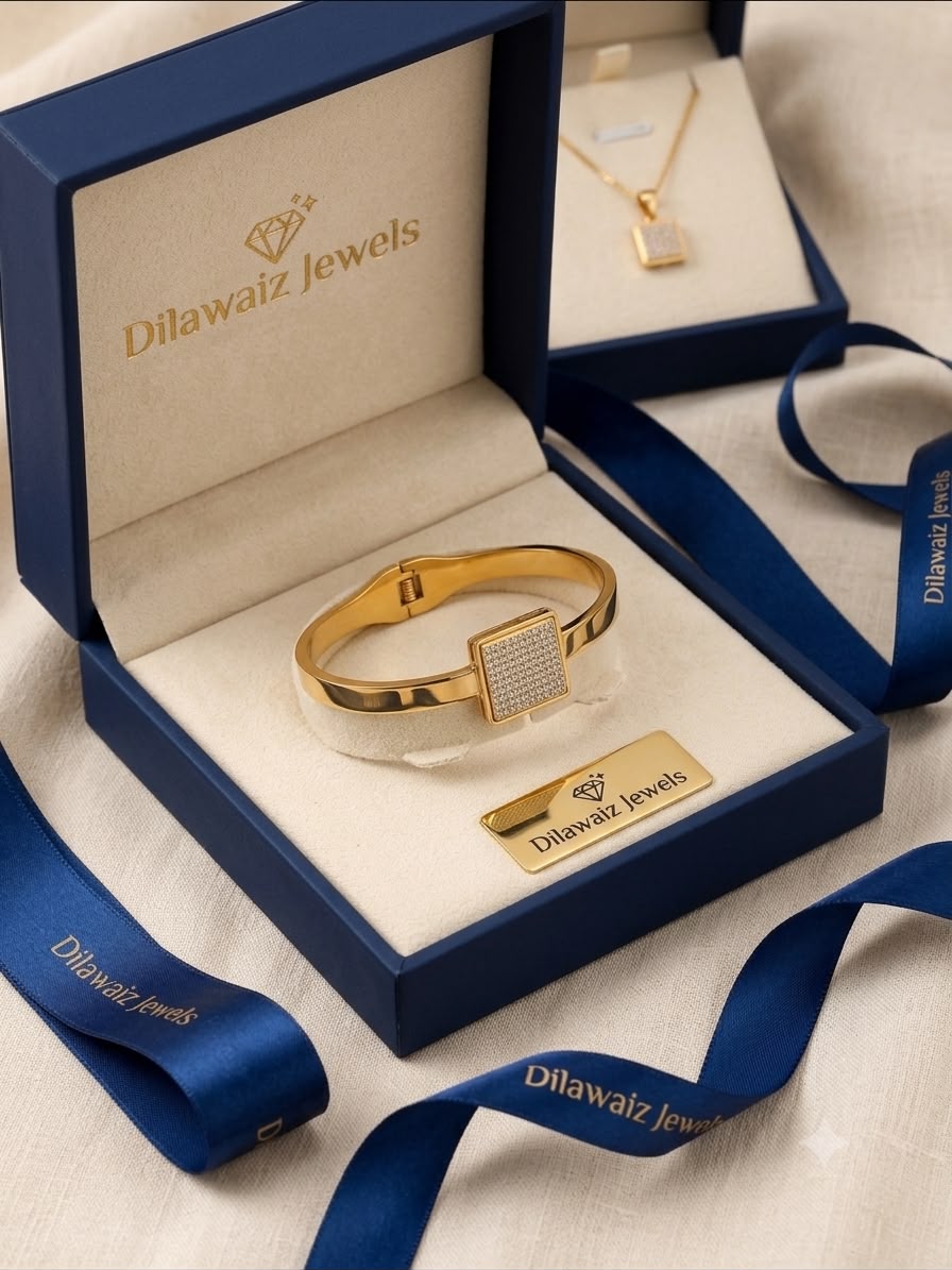 A gold bangle bracelet with a square crystal-embellished face, presented in an open navy blue Dilawaiz Jewels box. A matching gold necklace is displayed in a second box in the background, with blue branded ribbons draped around the boxes on a light beige fabric surface.