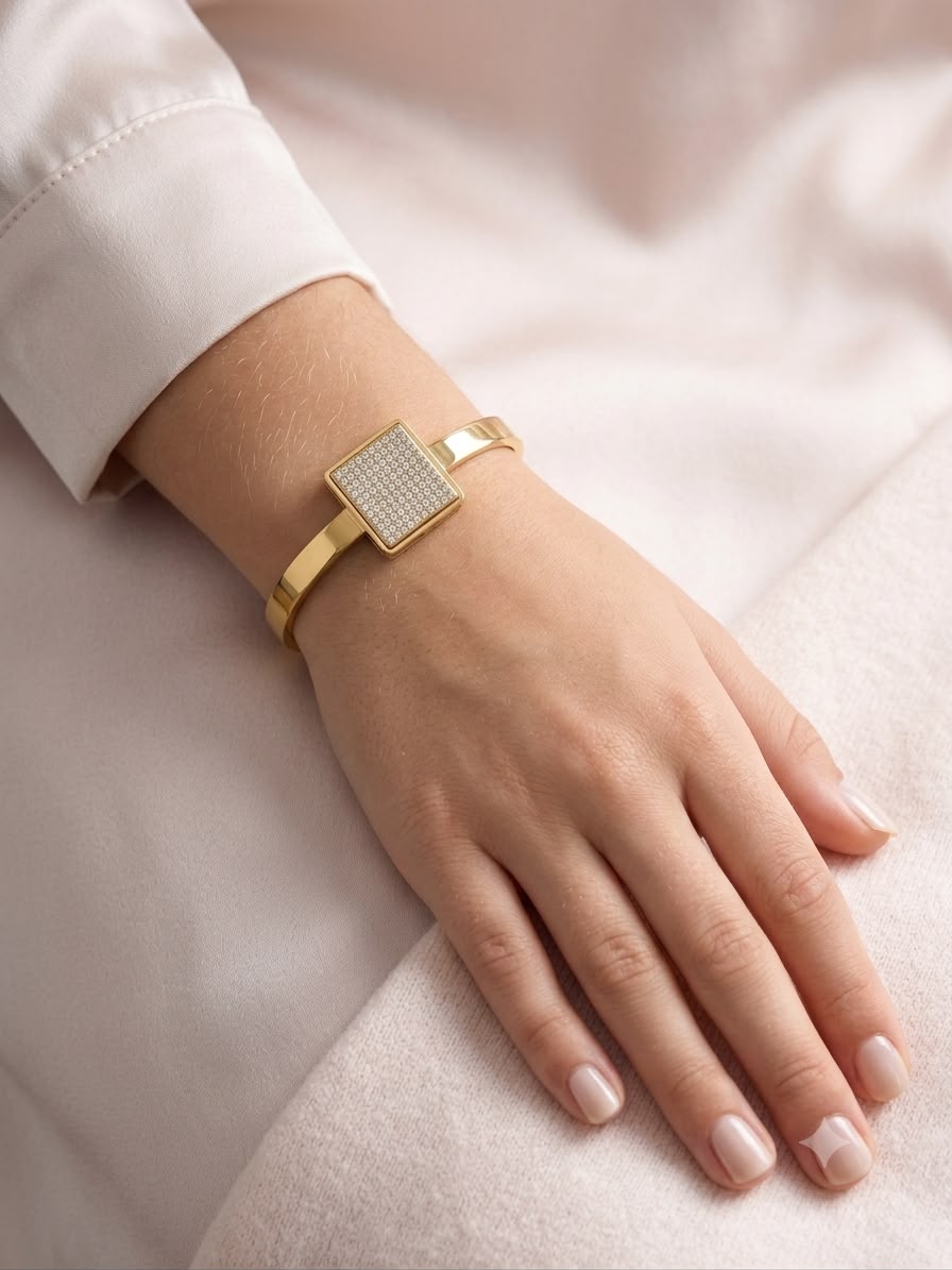 A gold bangle bracelet with a square crystal-encrusted face, shown worn on a woman's wrist against a soft pink fabric background.