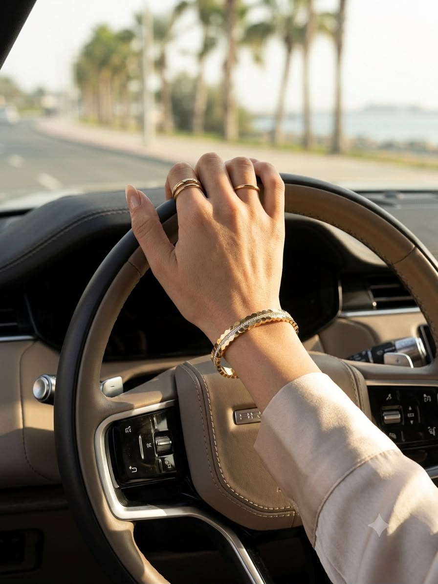 A close-up lifestyle shot of a person driving a car, wearing a gold chain bracelet on their wrist and gold rings on their fingers. The steering wheel is beige leather, and the background shows a blurred road lined with palm trees and water.