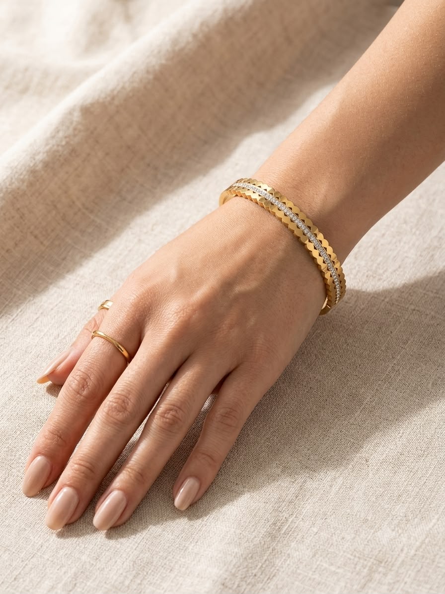 A close-up of a person's hand and wrist wearing two gold bangle bracelets, one with a textured honeycomb pattern and the other with a smooth snake-oil finish, resting on a beige linen fabric background.
