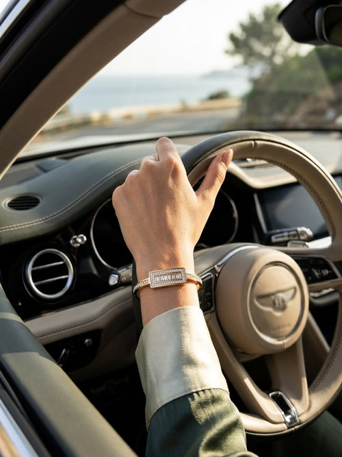 A close-up lifestyle shot of a person's hand resting on a car steering wheel, wearing a rectangular silver-toned metal bracelet with a warm gold-toned band and a small central crystal detail.