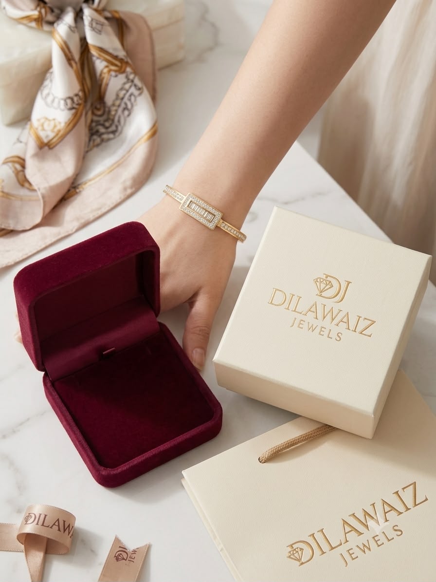 A close-up of a person's hand wearing a delicate gold-toned diamond bracelet, next to an open deep burgundy velvet jewelry box, a cream-colored Dilawaiz Jewels branded box, and a shopping bag on a light marble surface.
