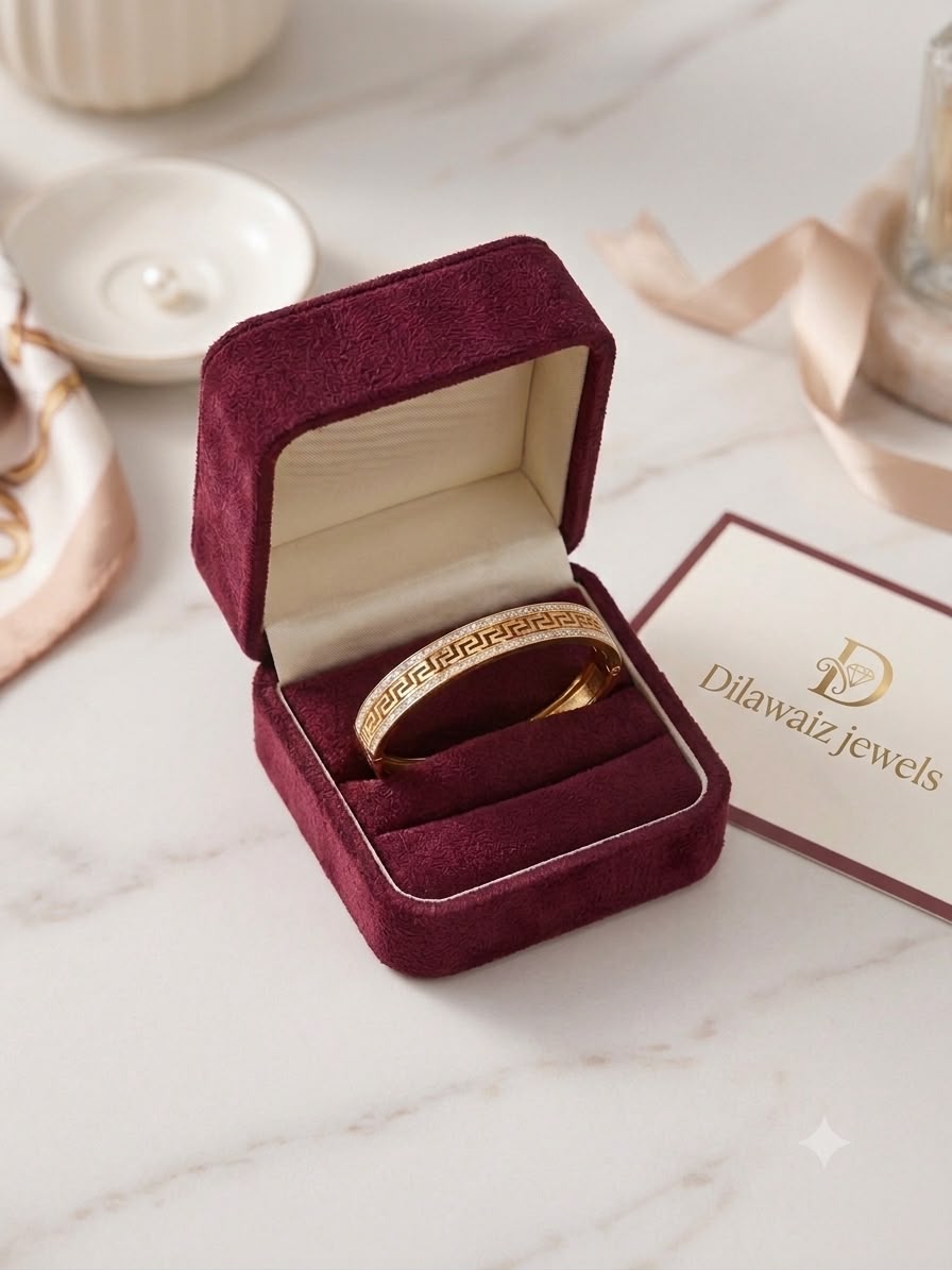 A gold band ring with Greek key engraving displayed inside an open burgundy velvet jewelry box on a white marble surface, with a Dilawaiz Jewels card nearby.