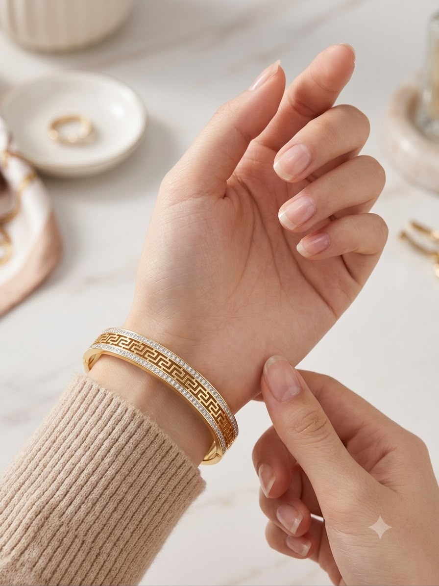 A close-up lifestyle shot of a person's wrist wearing a gold bangle bracelet with a Greek key pattern and small clear stones, paired with a beige ribbed sweater. The background is a soft-focus marble surface with jewelry props.