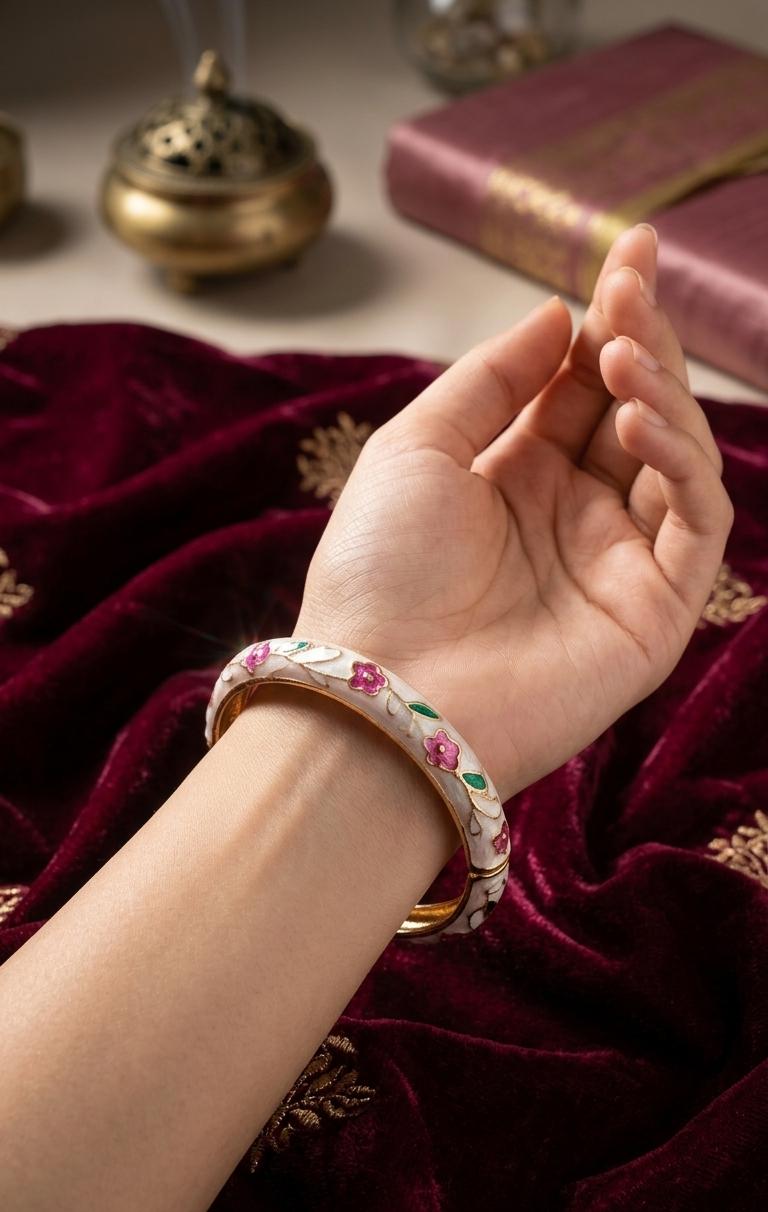 A close-up of a hand wearing a white bangle bracelet decorated with pink and green floral enameled details, resting on a deep red velvet fabric with gold embroidery.