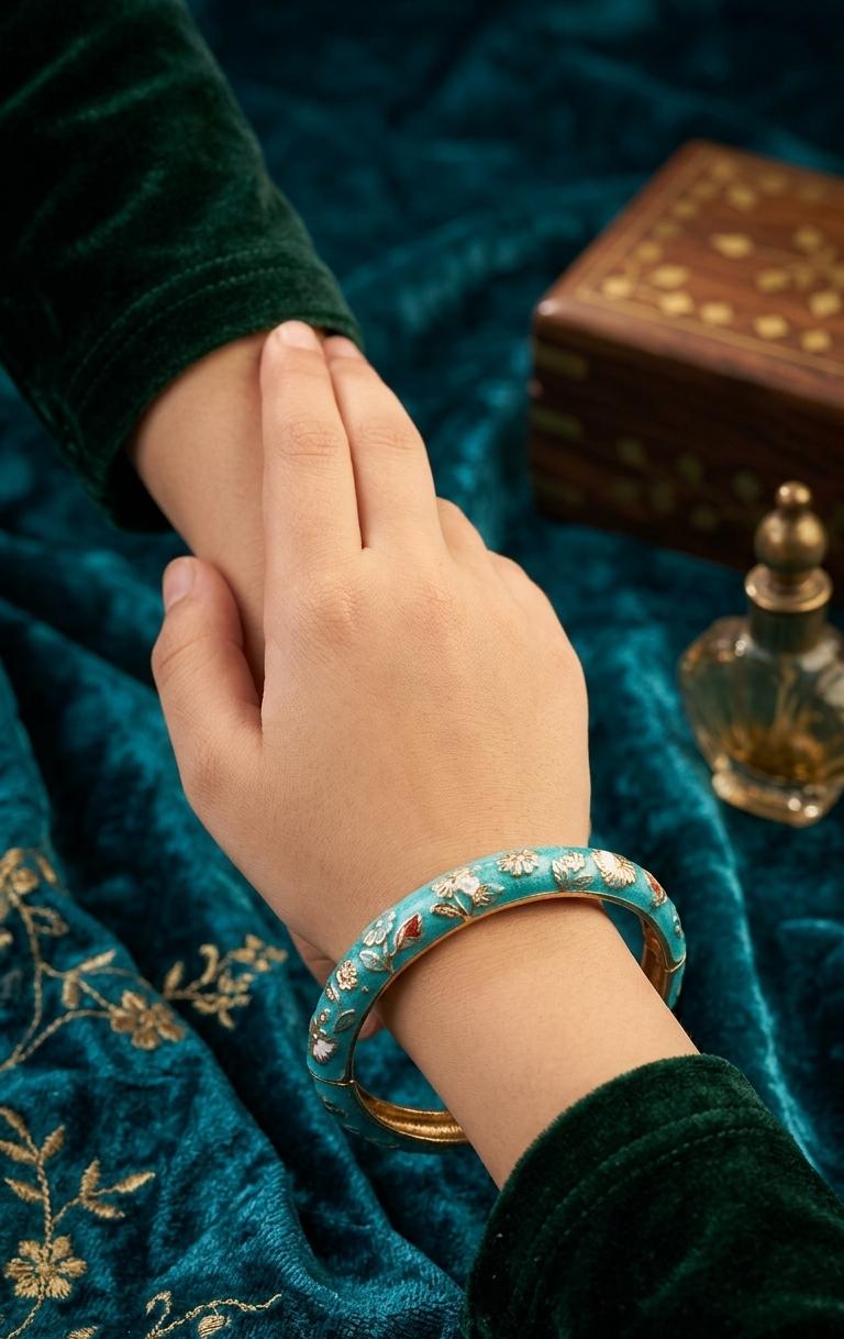 A close-up of a hand wearing a turquoise and gold floral-patterned bangle, resting on a dark teal velvet fabric with gold embroidery, alongside a dark green velvet sleeve and a decorative wooden box.