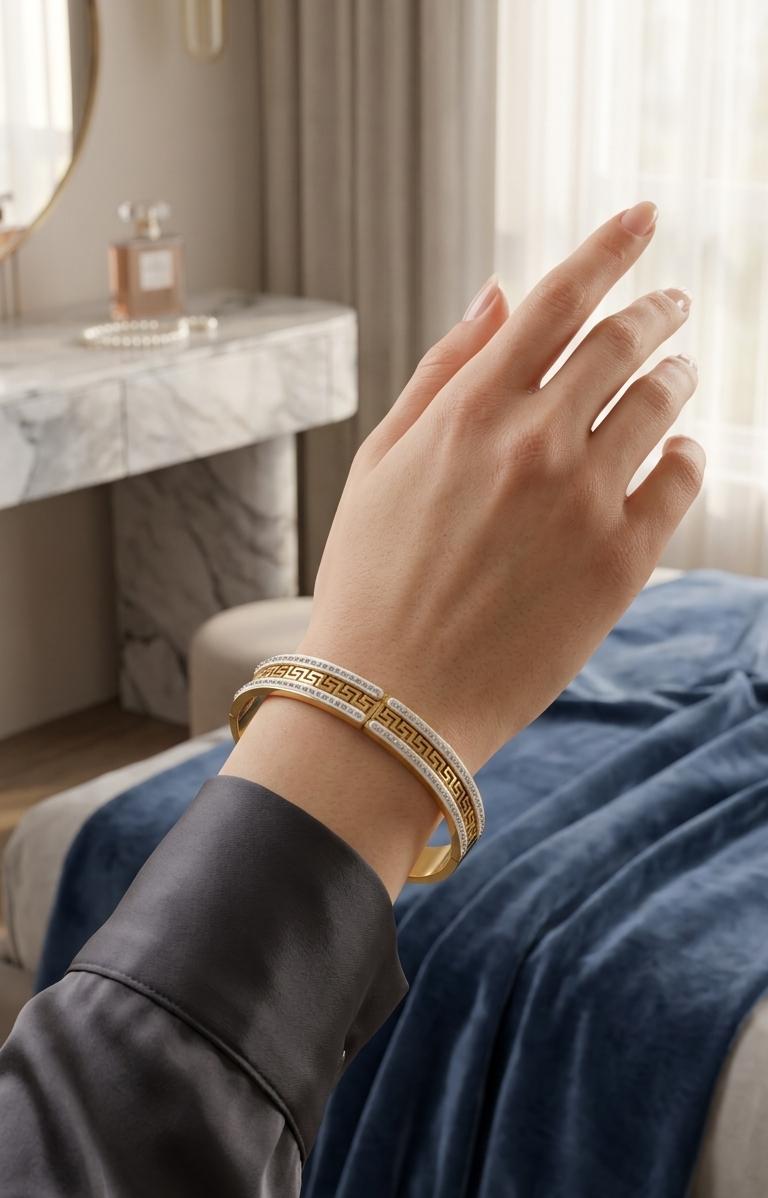 A close-up of a person's hand wearing a gold bangle bracelet with white inlay, set against a softly blurred bedroom background with a blue blanket and window curtains.