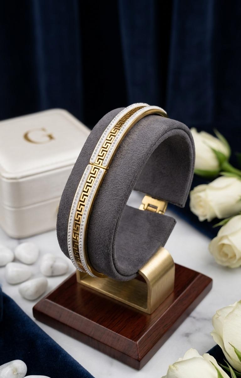 A gold and white beaded cuff bracelet displayed on a gray velvet ring stand with a wooden base, styled with white roses and a white jewelry box on a marble surface.